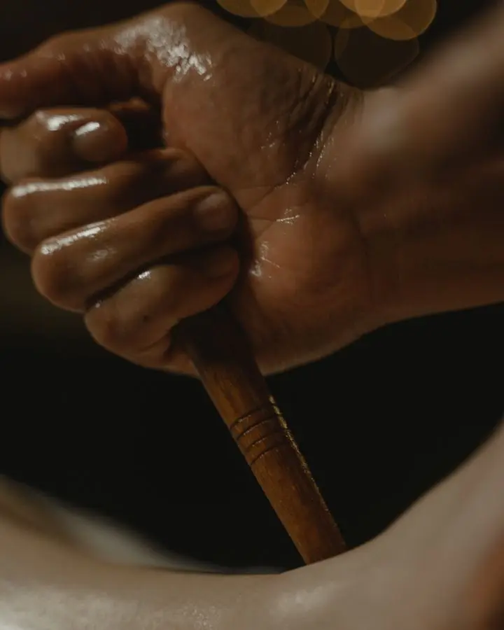 A close-up of a hand holding a wooden massage tool, set against a dark background.