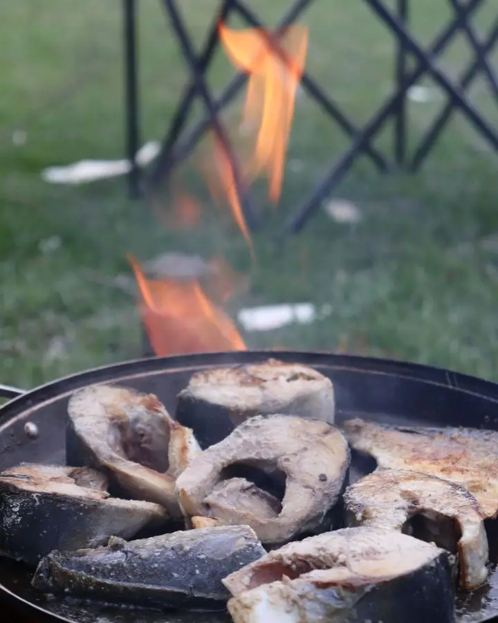 Slices of fish grilling on an outdoor flame, surrounded by nature.
