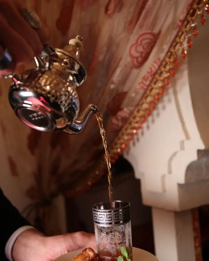 Elegant hand pouring tea into decorative glass with sweets on plate, ornate setting.